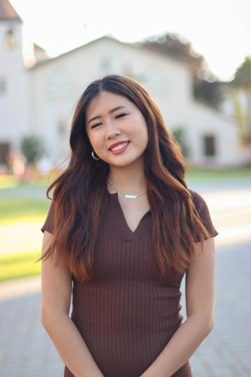 A person smiling outdoors with buildings in the background.