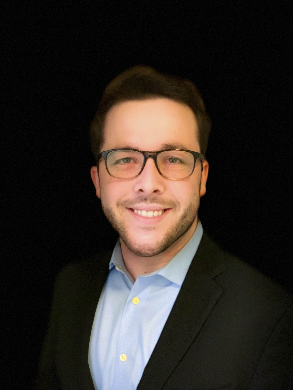 A person wearing glasses, a suit, and smiling against a black background.