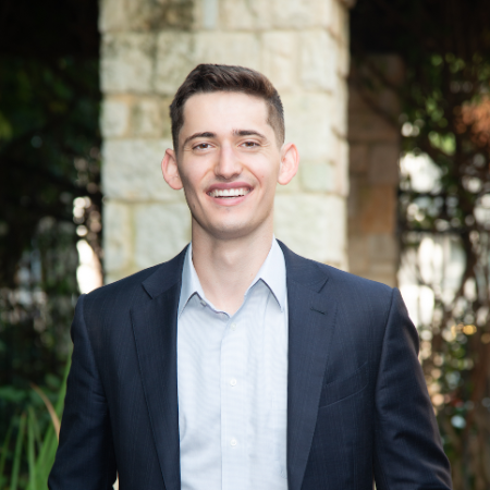 A person smiling, wearing a blazer in an outdoor setting.