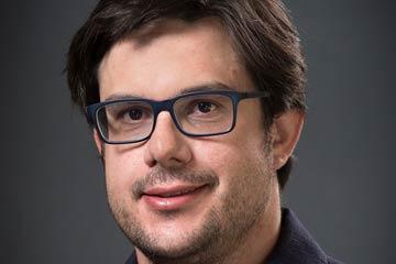 A person with glasses smiling against a dark background.