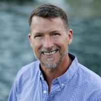A person standing outdoors near water, wearing a blue shirt.