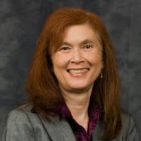 A woman in professional attire, smiling against a dark background.