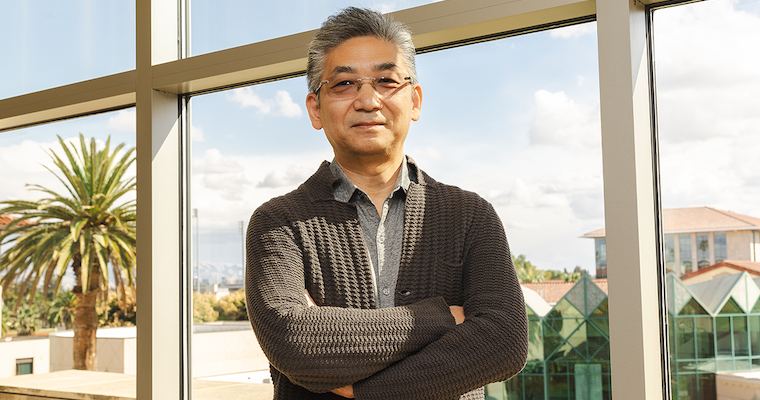 A Korean man in tinted glasses and a brown cardigan stands, arms crossed, in front of a window, palm tree and SCU campus behind him.