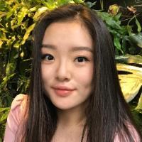 Young woman in a garden, wearing a light pink top.