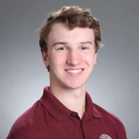 A person wearing a maroon shirt smiling against a gray background.