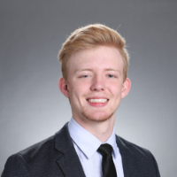 A smiling individual wearing a suit and tie with a grey background. 