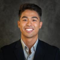 A smiling person in a suit against a dark background. 