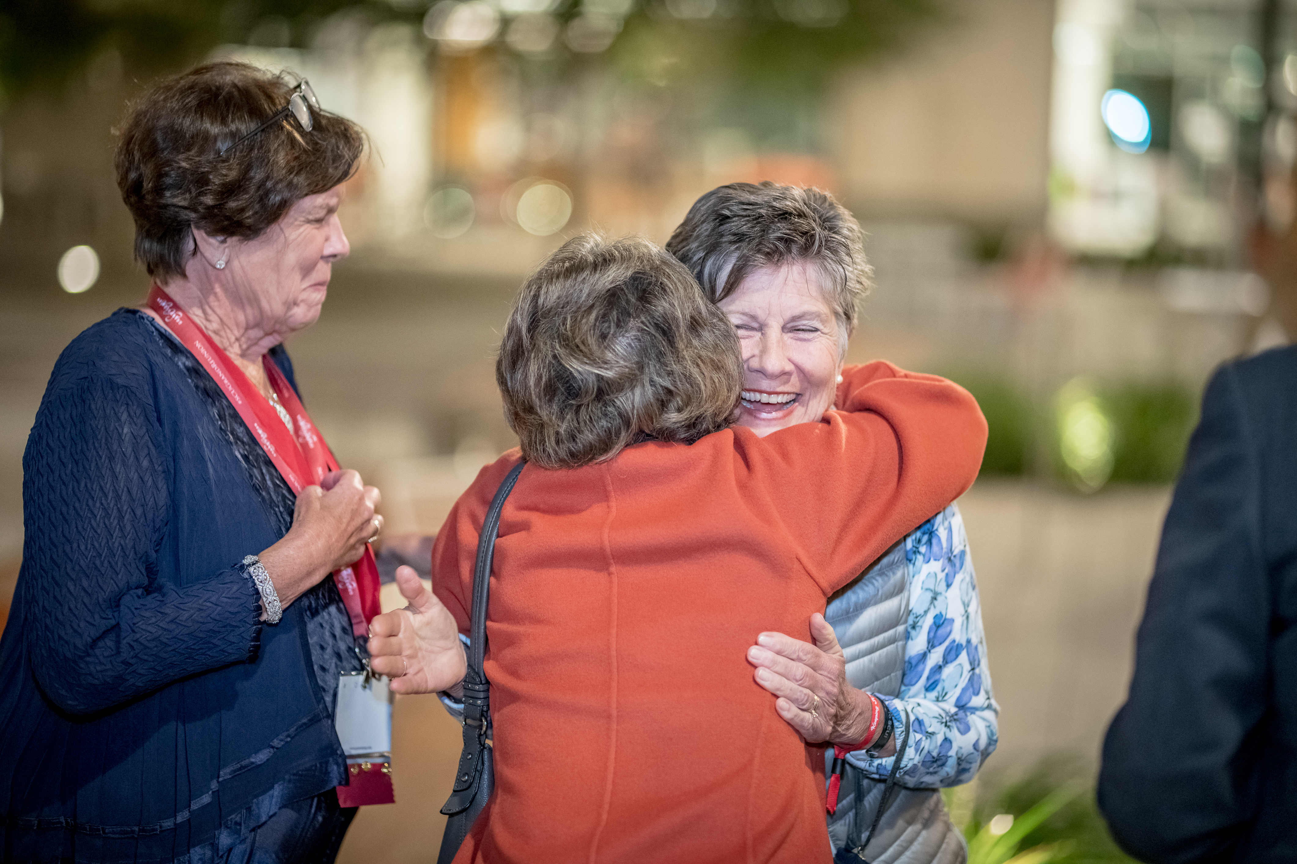 One woman smiles as two other women hug each other