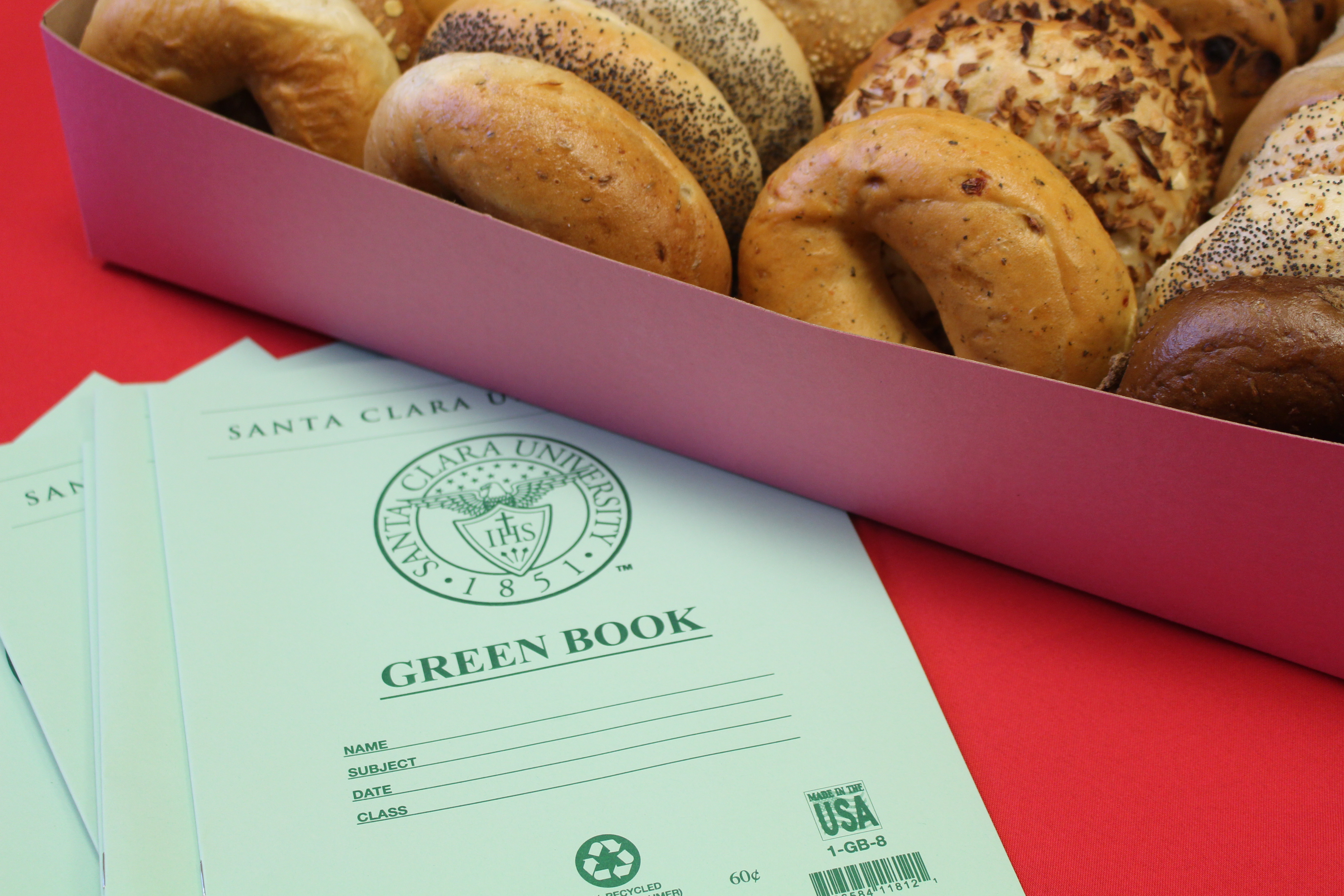 Alt text: Box of assorted bagels next to green notebooks on a red surface.