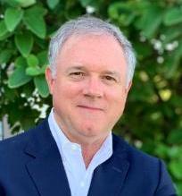 A man in a suit stands with arms crossed in front of greenery.