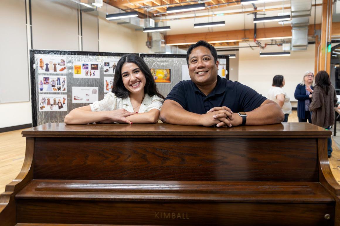 A smiling woman and man pose with their arms on top of a piano in a rehearsal room. Behind them other actors converse near a pair of bulletin boards full of costume and stage design images.