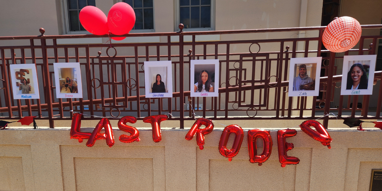 Pictures of several SCU Alumni Association student workers hung up on the metal rails of a walkway. Red and white decor surrounds the photos and red foil balloons hung up underneath the photos read 