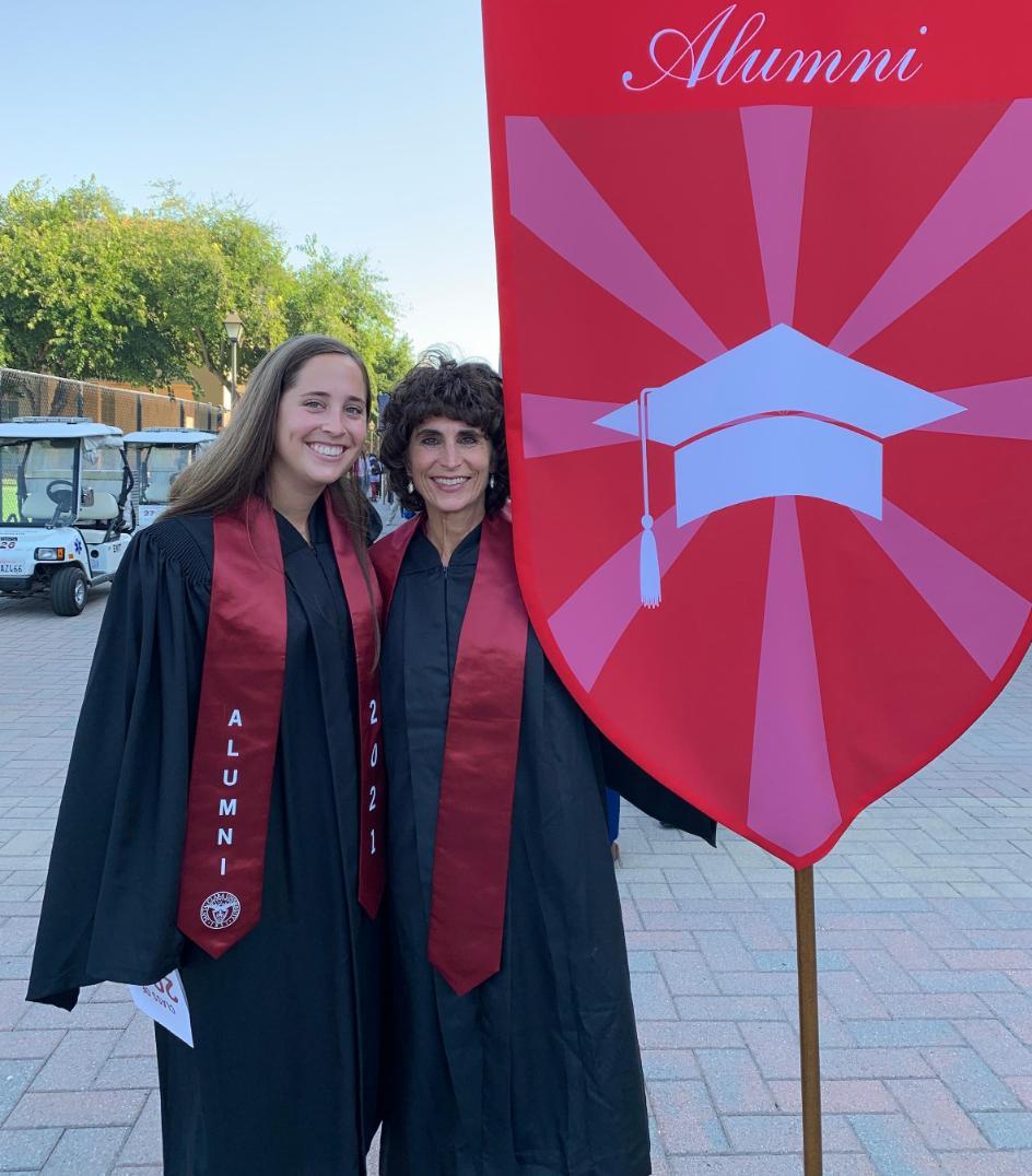 Two people in graduation gowns next to a ceremonial banner.