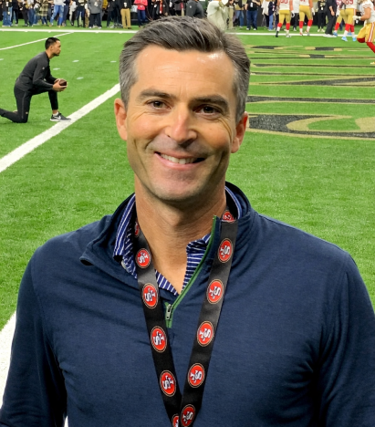 A smiling man wearing a San Francisco Giants lanyard and standing in front of a football field.