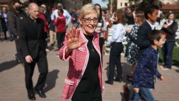 SCU President Julie Sullivan smiles and waves at the camera.