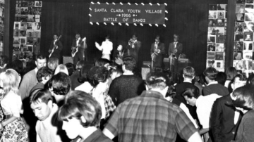 Young men and women in 1960s clothing dancing in front of a stage with a band. A sign over the band reads 