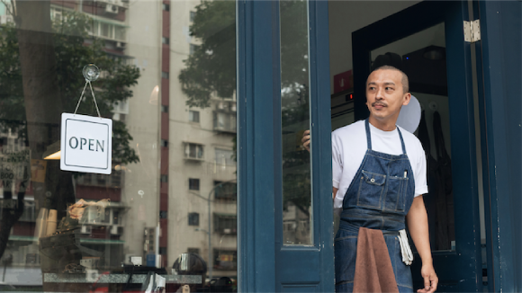 A man in a denim apron with a towel in the pocket looks out of a storefront. A sign in the window reads 