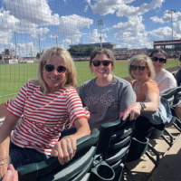Four smiling women sitting in stadium chairs in front of a baseball field