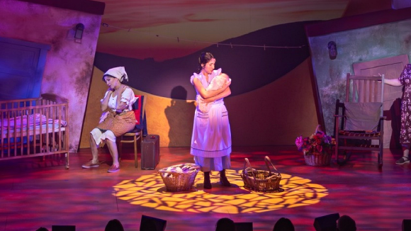 A young woman in an apron holding a baby, standing on a theatre stage.