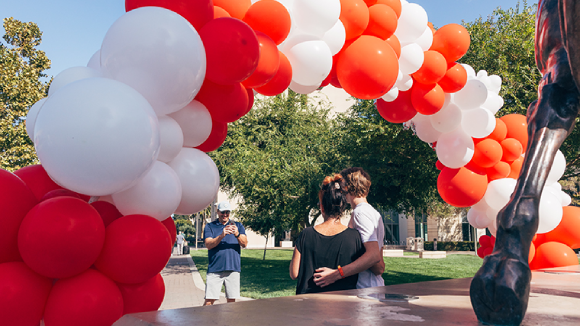 A man takes a picture of two people under a red and white balloon arch in front of a Bronco statue at Santa Clara University.