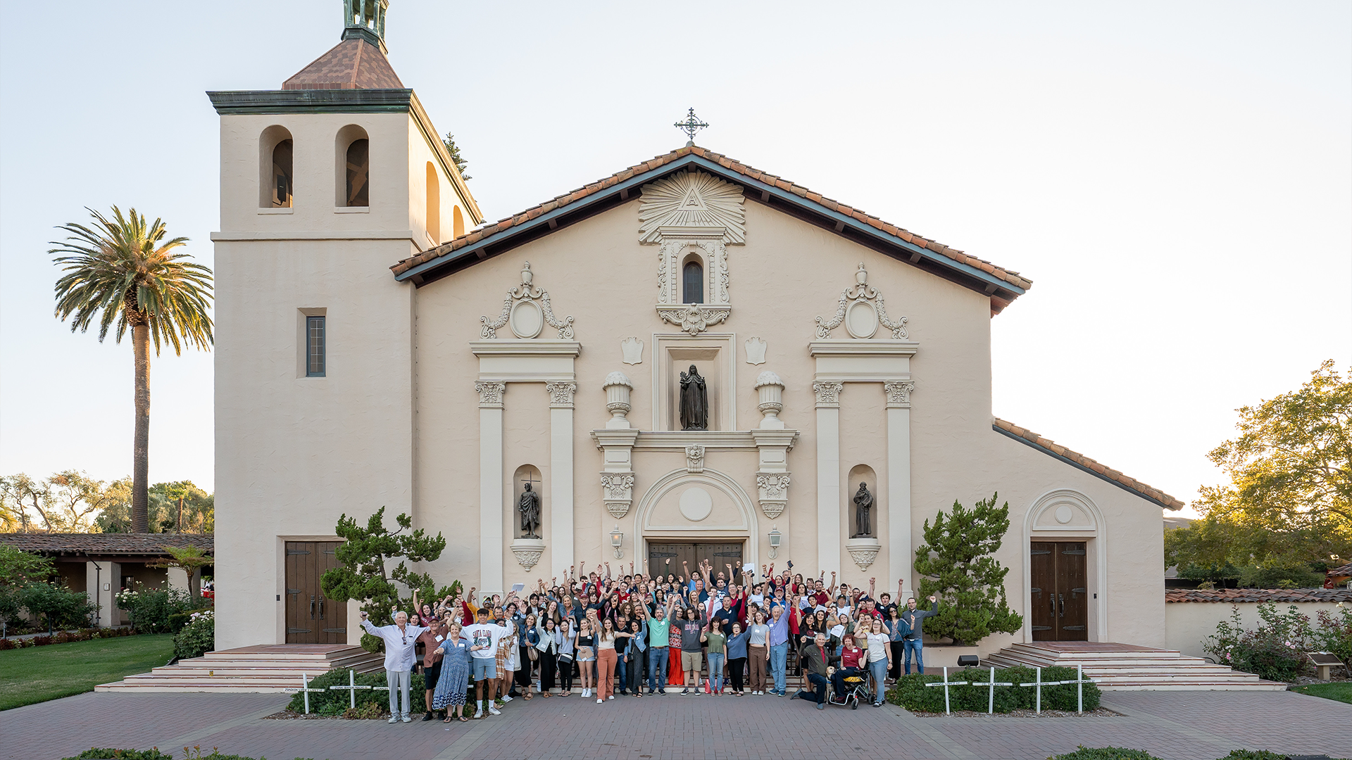 A large group of alumni wave at the camera in front of Mission Santa Clara