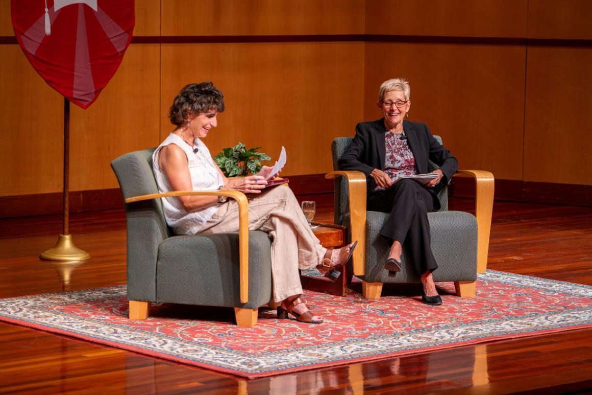 Two women seated for a formal chat. They are smiling and both have notes in hand.