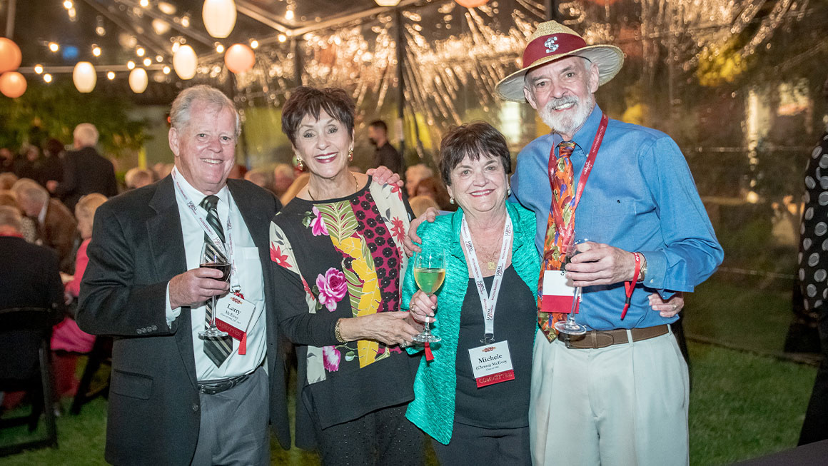Four alumni standing in front of a lit pavilion and smiling at the camera 