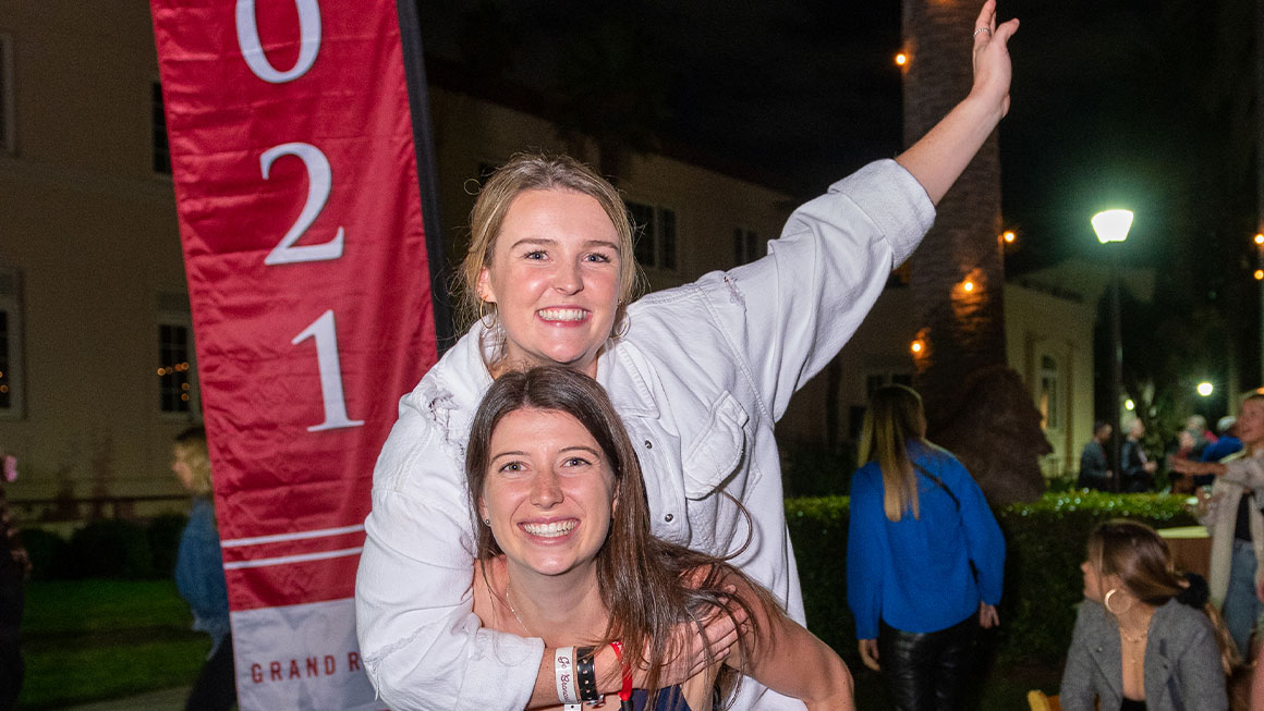 Two women cheering in front of a banner that reads Class of 2021 