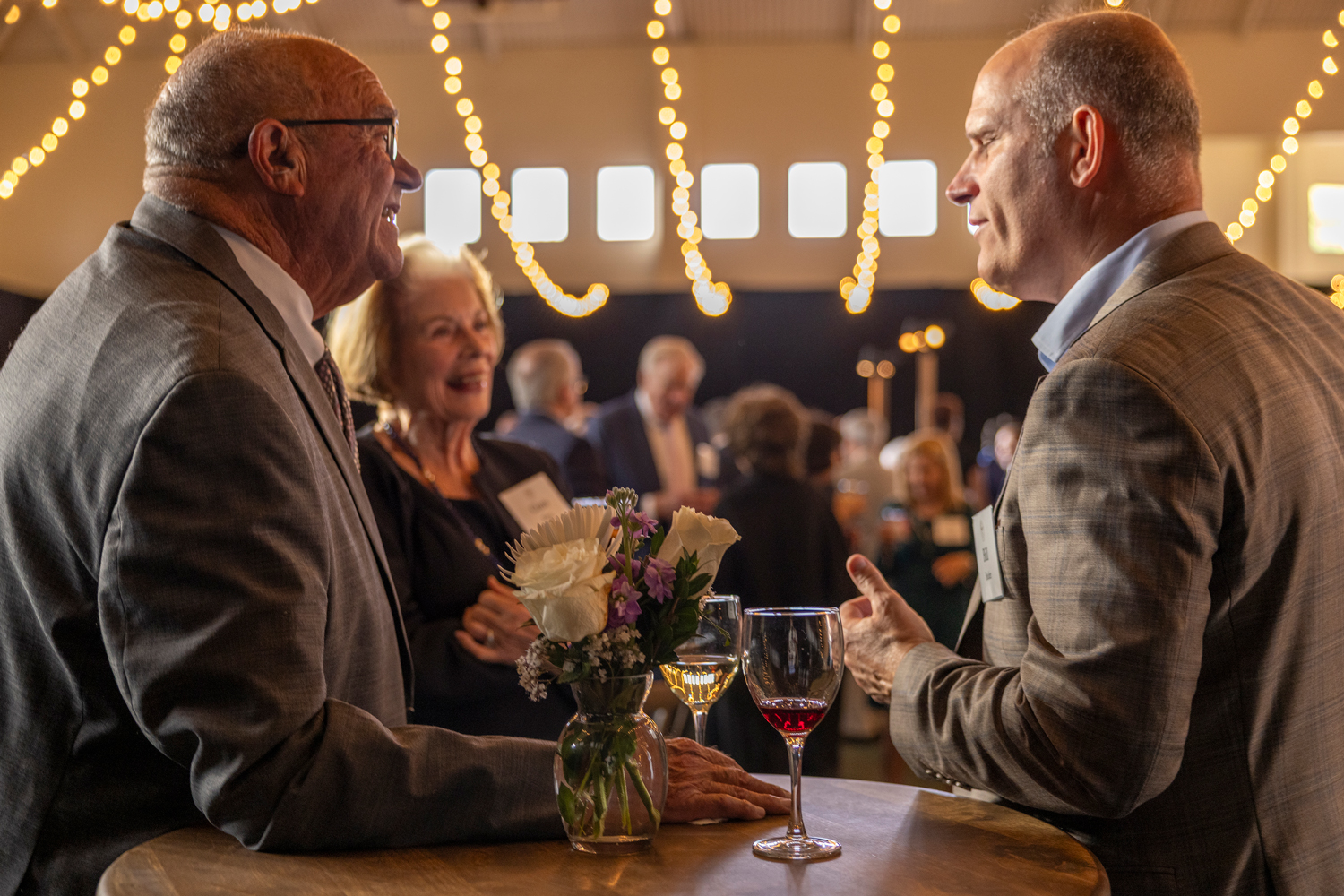 Two men talking with a large crowd in the background in a room lit by bistro lights