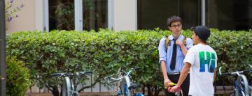 Two people standing and talking in front of a hedge.