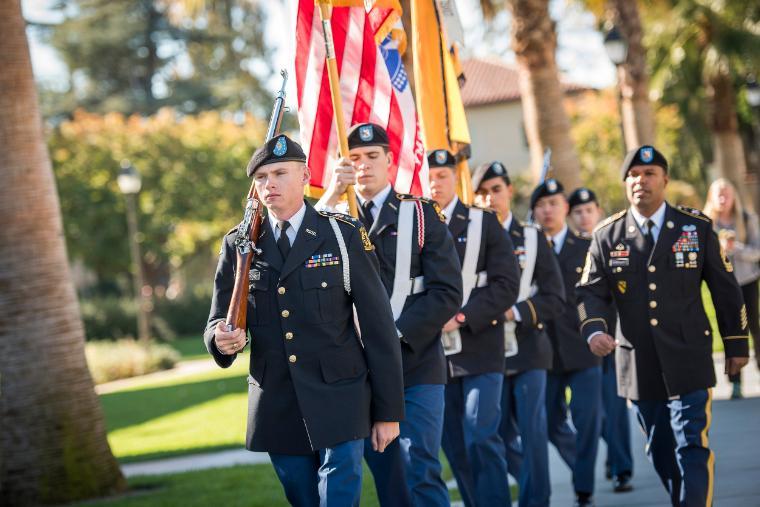 A group of veterans in uniform march, holding flags.
