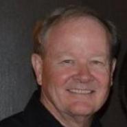 A person smiling, wearing a dark shirt against a dark background.