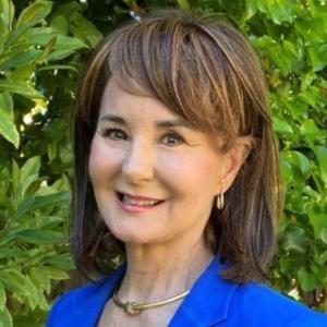 A person in a blue blazer stands in front of green foliage.
