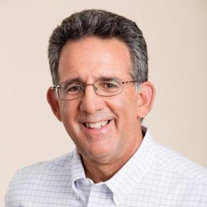 A person smiling, wearing glasses and a light-colored shirt.