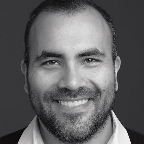 A man smiling, wearing a shirt and sweater, against a dark background.