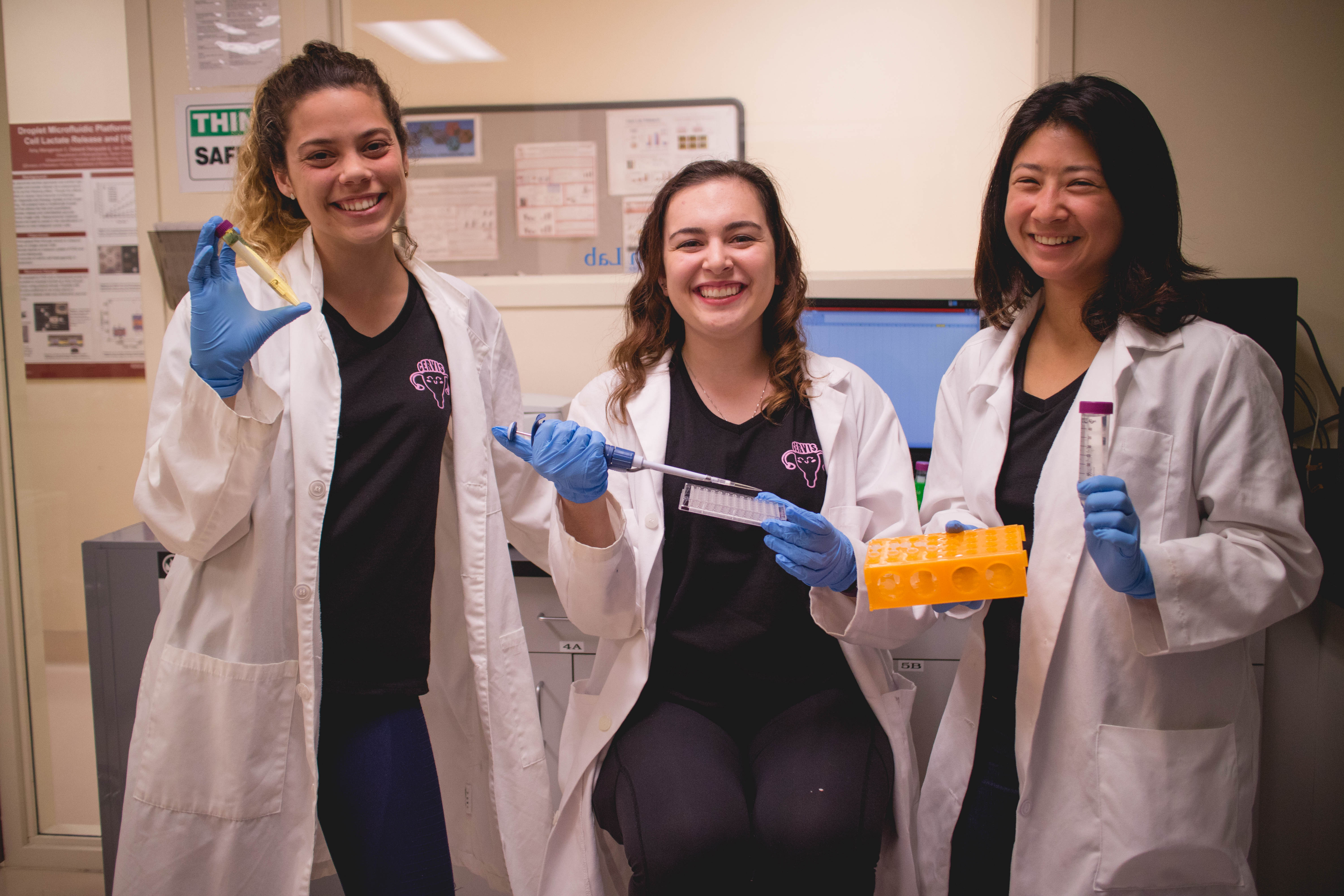 Three people in lab coats holding scientific equipment and smiling in a laboratory.