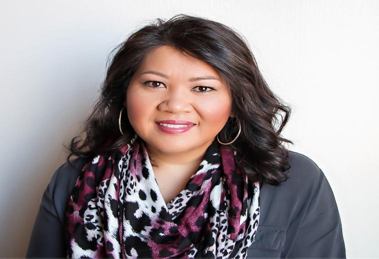 Alt text: Woman wearing a patterned scarf and grey shirt smiling in front of a plain background.
