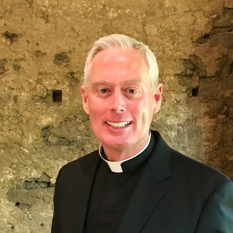A person wearing clerical clothing smiles in front of a textured stone wall. image link to story