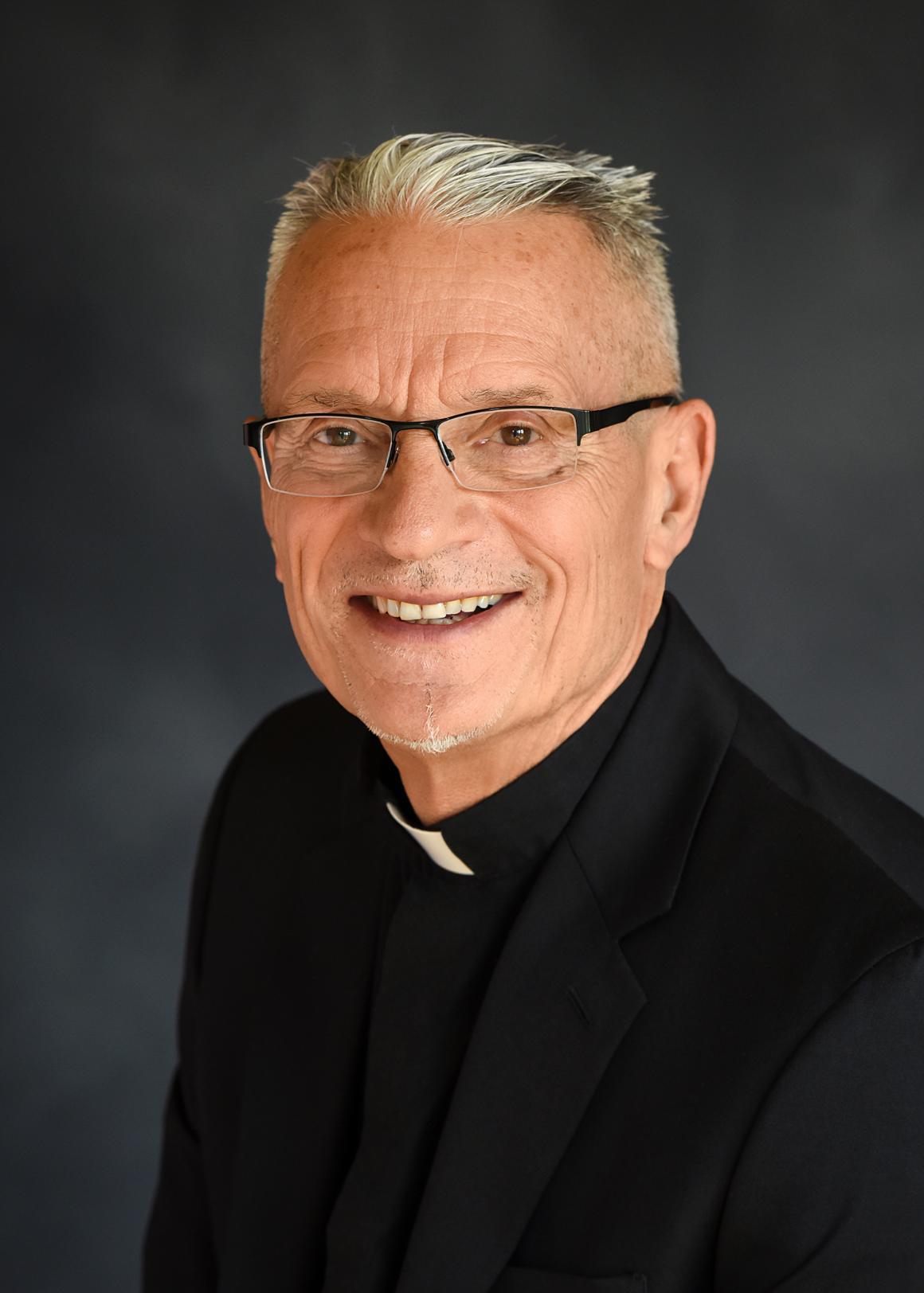 A smiling man in clerical attire with a dark background. image link to story