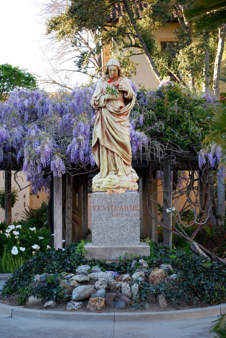 A garden with a statue of Jesus amidst purple wisteria flowers.