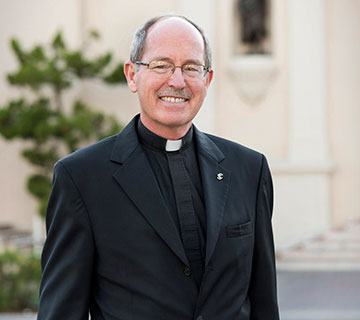A man in clerical attire standing outdoors, smiling.