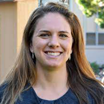 A person smiling outside with buildings and trees in the background. 