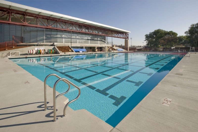 Outdoor swimming pool with clear blue water, pool lanes, and a modern building in the background.