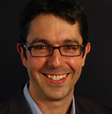 A person wearing glasses and smiling against a black background. image link to story