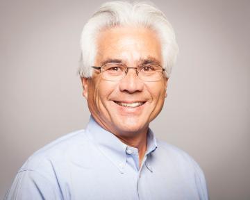 A man smiling, wearing glasses and a light blue shirt.