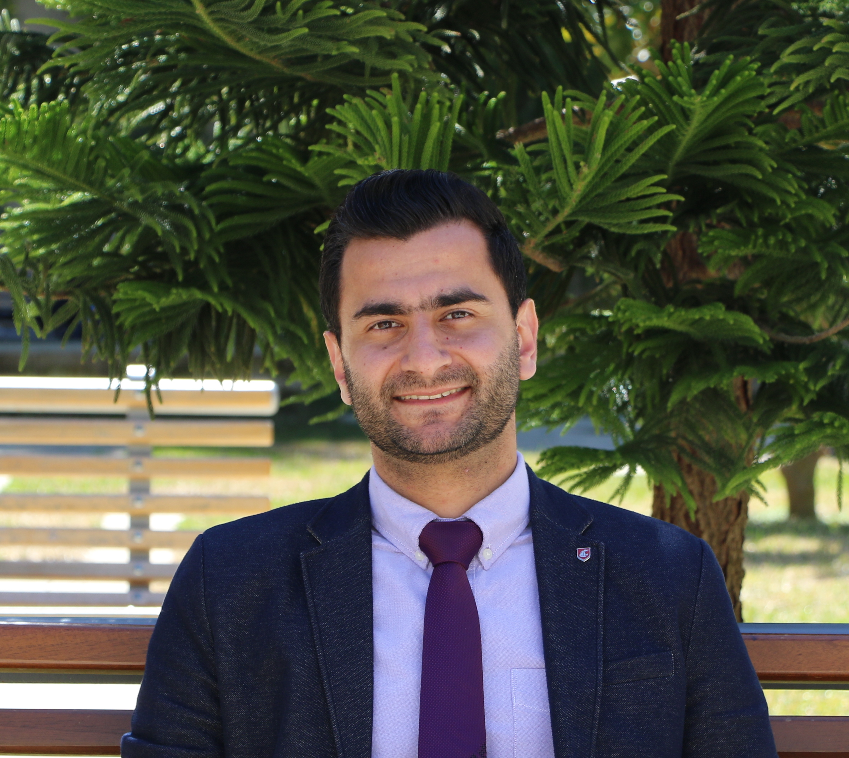 A man in a suit standing outdoors with trees in the background.