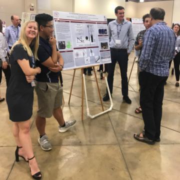 Female student smiles at the camera while male teammate talks about their poster with visitors image link to story
