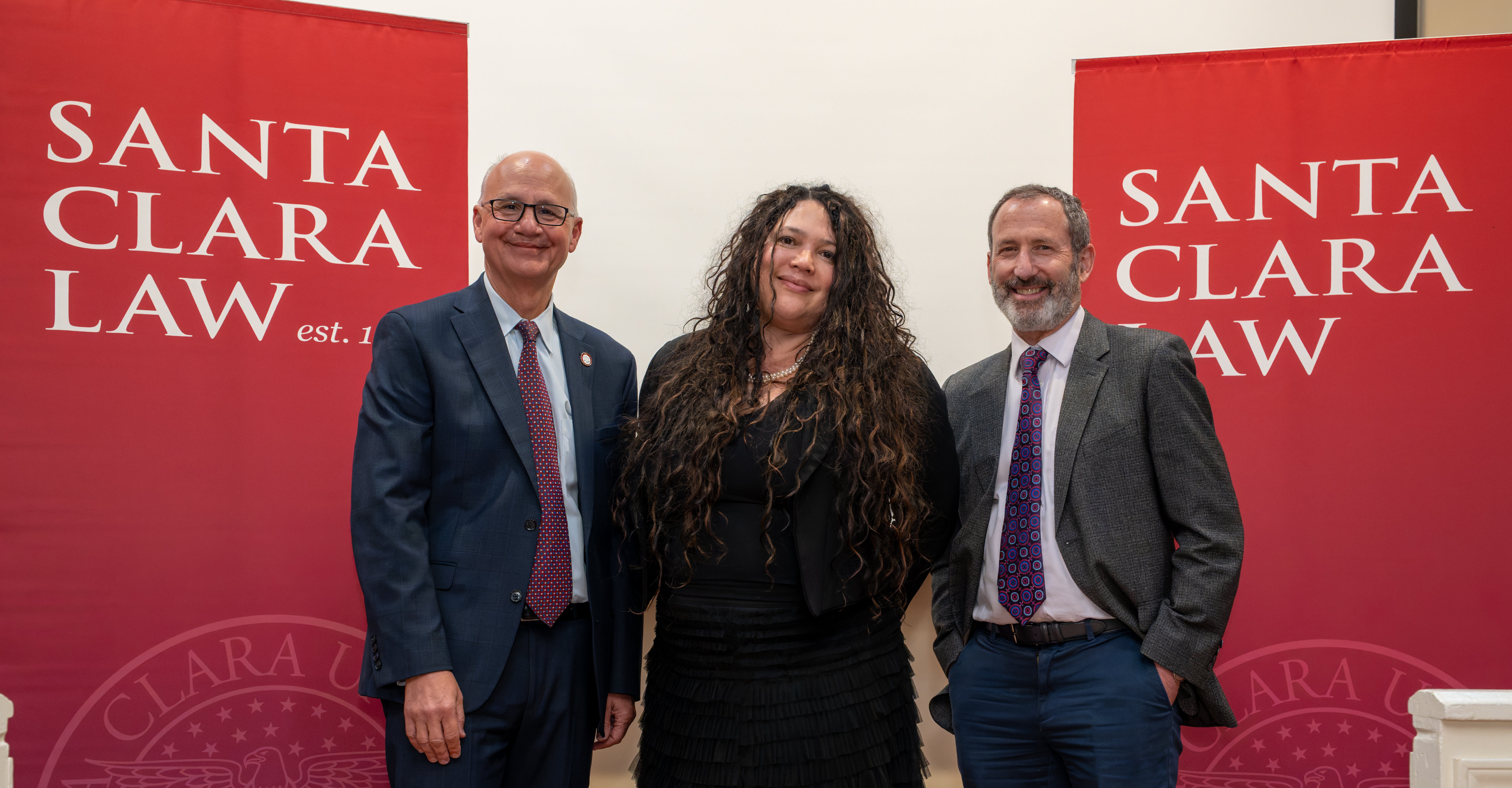 Three people stand in front of red Santa Clara Law banners, smiling and posing for a photo.
