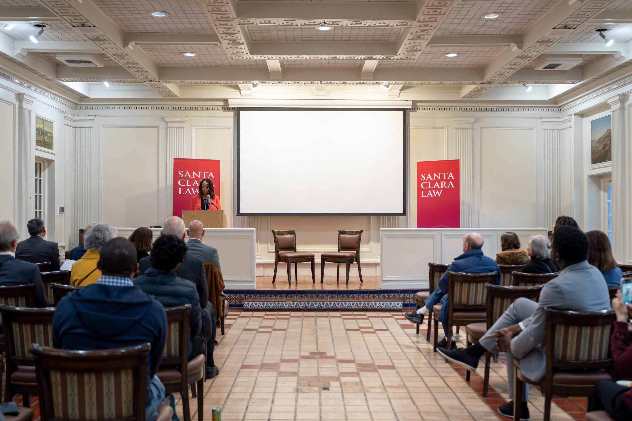 A speaker at a podium addresses an audience in a formal Santa Clara Law event hall.