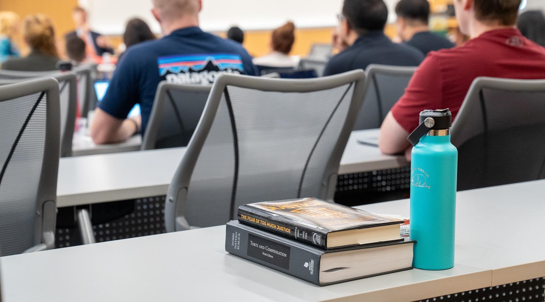 two books stack on top of each other next to a teal color metal water bottle on a desk in a classroom filled with students watching a speaker talk at an event 